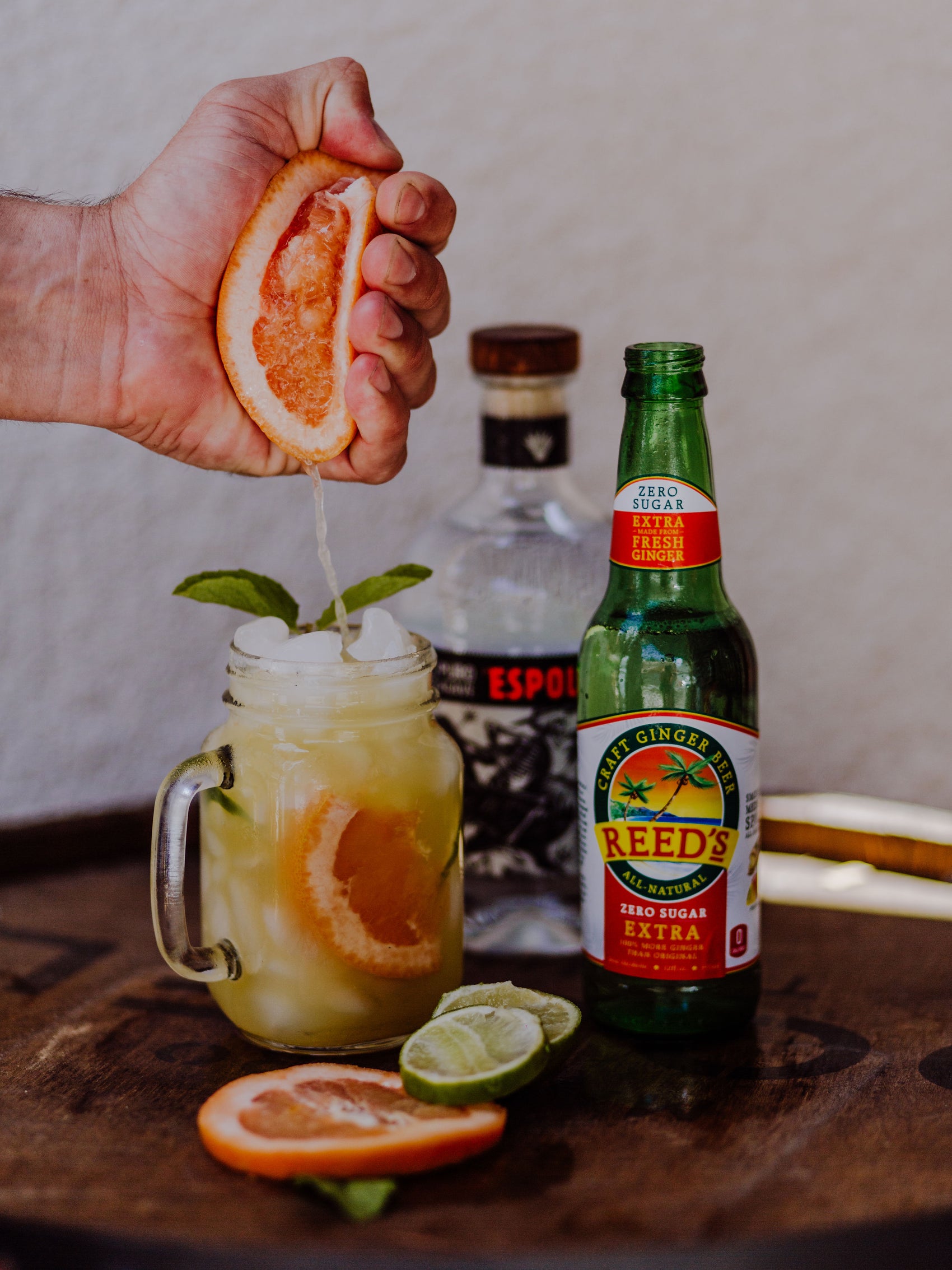 Someone squeezing an orange into a glass next to a glass bottle of Reed's Zero Sugar Extra Ginger Beer.