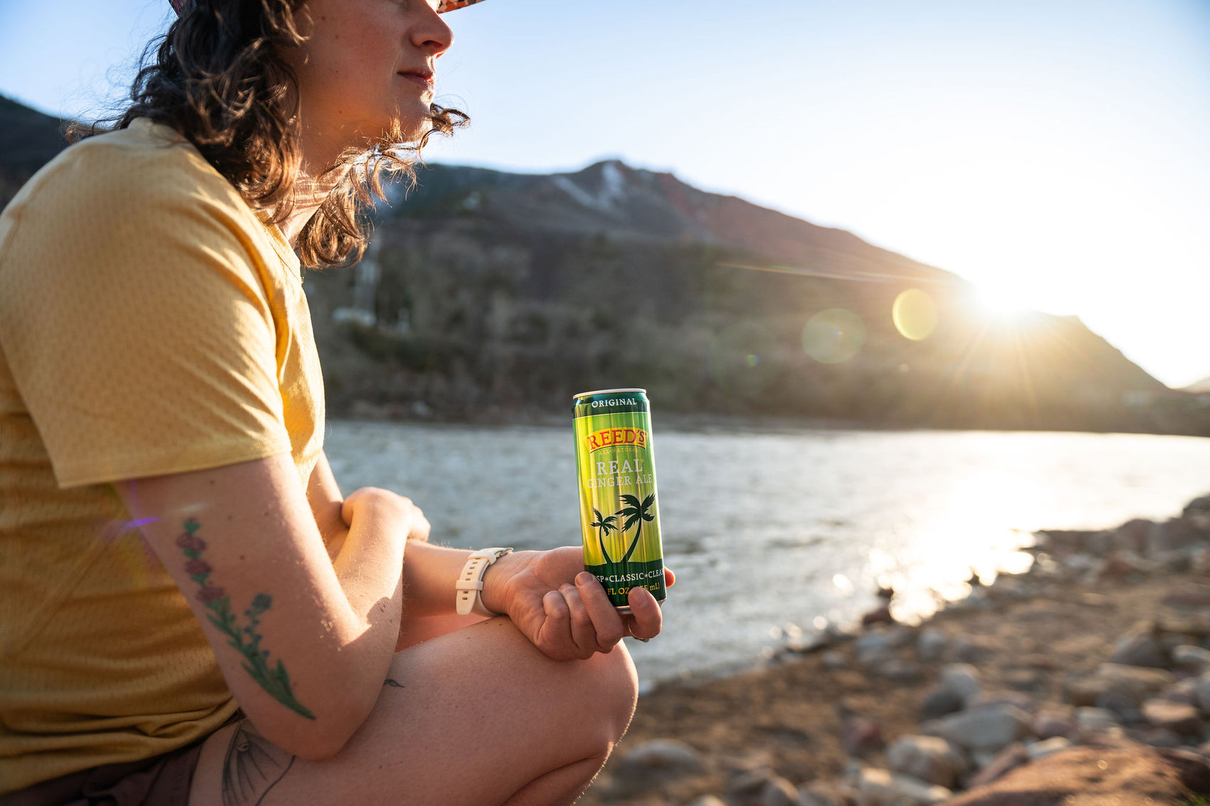 A person wearing a yellow shirt holding a can of Reed's Real Ginger Ale in front of a body of water.