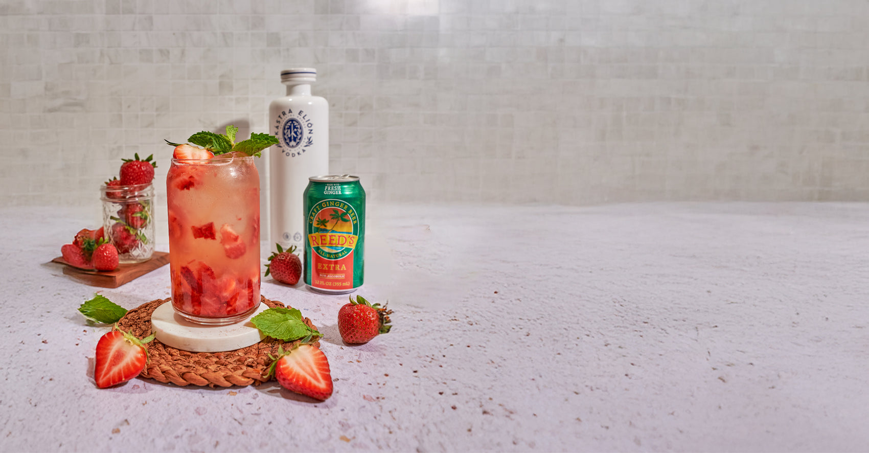 A pink drink in a glass garnished with mint and strawberries on a brown coaster next to a can of Reed's Extra Ginger Beer on a white counter.